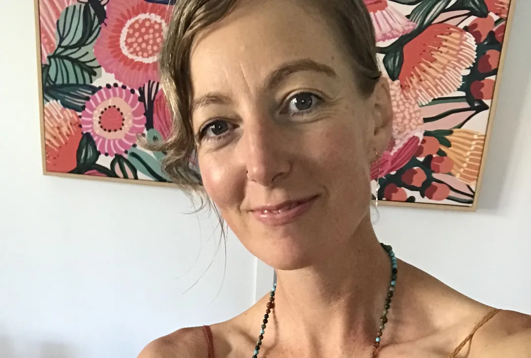 A smiling person with dark blonde hair and kind eyes.  Their hair is pulled back and fringe sweeps to one side in light waves.  They are wearing a colourful beaded necklace and stand in front of a colourful abstract picture of native Australian flowers in pinks, oranges and greens.