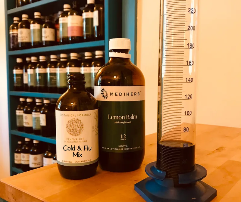 In the foreground are two amber bottles on a wooden bench, beside a large measuring cylinder.  One bottle is a herbal tincture labelled 