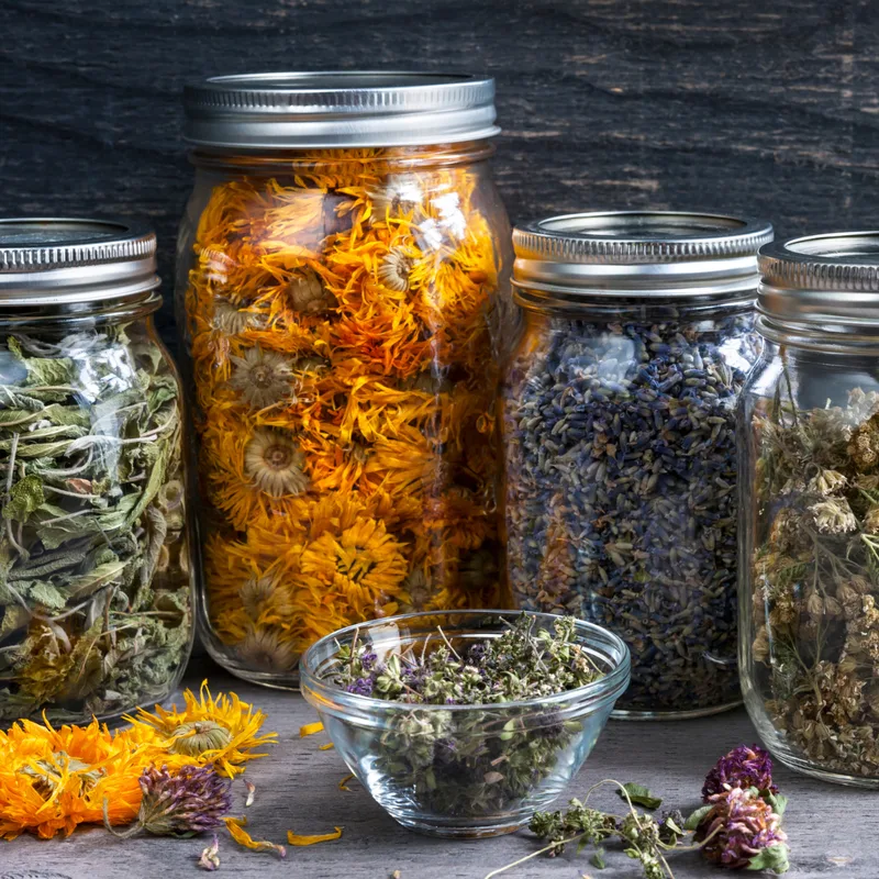 Jars filled with dried herbs and flowers, including calendula and lavender, sit on a wooden surface. A small glass bowl of dried herbs is in the foreground.